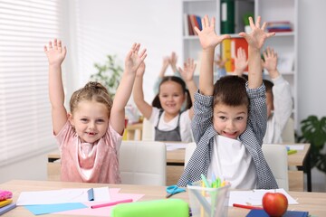 Happy children raising hands at wooden desks during art lesson in elementary school