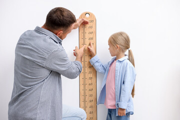 Father measuring his little daughter’s height with scale near white wall indoors
