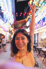 Woman smiling and raising arm, enjoying a night out in a vibrant hanoi street filled with colorful neon lights, capturing a happy moment during her travels
