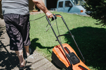 Obraz premium A person in casual clothing pushes an orange lawn mower across a sunny yard. Green grass and a driveway border the scene, with a blue van visible in the background.