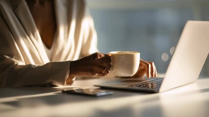 Woman in elegant beige blazer enjoying morning coffee while working on laptop at bright modern workspace. Productive start to day with hot beverage, digital technology in cozy office environment