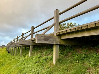Obraz premium Low-angle view of a wooden footbridge with railings standing on grassy ground under a cloudy sky.