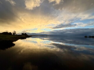 Obraz premium Calm lake at sunset with dramatic golden clouds reflected in the still water, creating a mirror-like landscape view