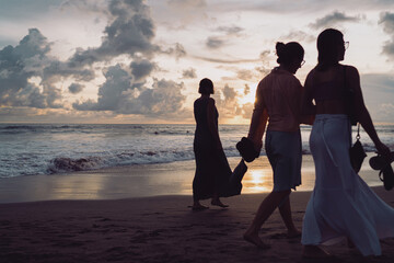 Group of women strolls barefoot along evening shore, dresses and skirts catching breeze as waves glow. Concept of friendship, unhurried conversation, and time well spent beside the sea