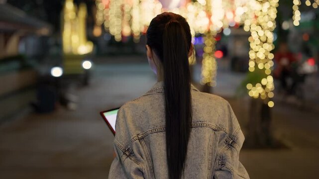 Asian woman with long black ponytail holds digital tablet and walks along night street rear view. Young Japanese lady with mobile device strolls past glowing trees in night town