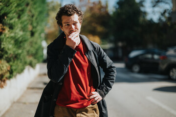 A man wearing a dark coat and red sweater stands outside on a sunny day, appearing deep in thought, with trees and cars creating an urban background.