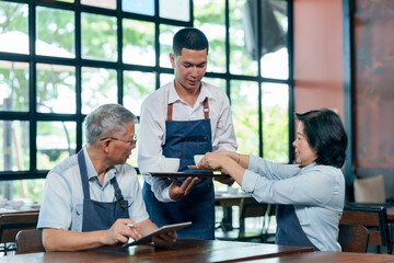 Senior asian couple restaurant owners wearing apron sitting at table instructing young male waiter...