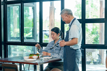 Asian elderly male owner standing beside female customer at table in small cafe, providing service chatting, friendly interaction, restaurant communication, teamwork in family small business