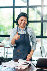 Asian elderly woman cafe owner wearing apron showing thumbs up with cheerful smile representing...