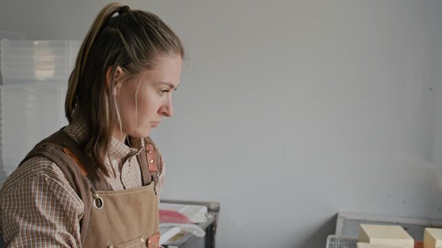 Tilt shot of skilled young Caucasian female cheesemaker in apron putting on latex gloves preparing to work with artisan cheese ripening on draining mat in cheese shop