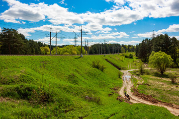 Fototapeta premium Green railway embankment and tracks stretch across a verdant landscape under a bright sky. Power lines connect infrastructure with nature, a dirt trail winding below, ready for adventure