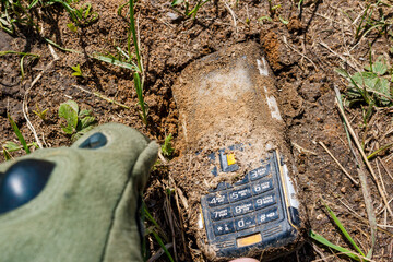 Grubby old cell phone unearthed from soil, keys caked with mud. A gloved hand reaching to retrieve...