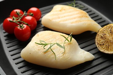 Pieces of tasty grilled squid, tomatoes, lemon and rosemary on black pan, closeup