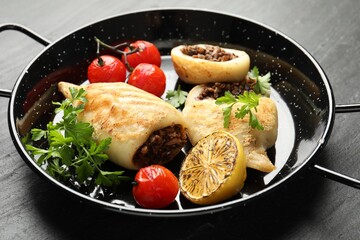Pieces of tasty grilled squid stuffed with mushrooms, parsley, tomatoes and lemon in pan on dark textured table, closeup