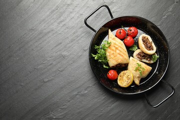 Pieces of tasty grilled squid stuffed with mushrooms, parsley, tomatoes and lemon in pan on dark textured table, top view. Space for text