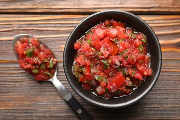 Tasty salsa sauce in bowl and spoon on wooden table, top view