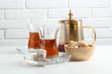 Tasty Turkish tea in glass cups, brown sugar, sweets and teapot on white marble table against brick wall, closeup