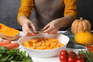 Woman adding thyme to pumpkin in baking dish at white wooden table, closeup