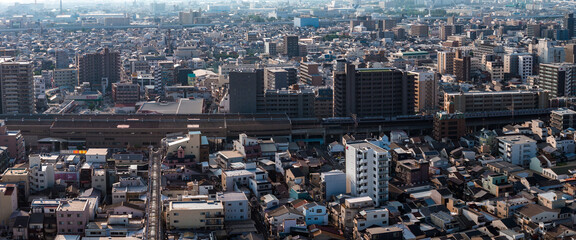 Obraz premium Aerial view of Osaka, Japan, shows mid rise apartments, tight houses, and an elevated railway with a passing train in hazy afternoon light across a vast district.