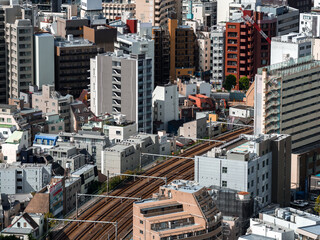 Fototapeta premium Aerial view of a dense Tokyo district with mid rise apartments and offices, parallel railway tracks curving between blocks, a red cylindrical tower, and sharp afternoon light.