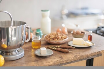 A bright kitchen counter in the morning showcases ingredients for baking. A stand mixer sits next to bowls of nuts, eggs, butter, and a bottle of milk, ready for use