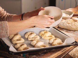 Woman stands in kitchen, carefully brushing egg wash onto twisted bread dough on baking sheet. Background shows flour, walnuts, and more dough, ready for baking