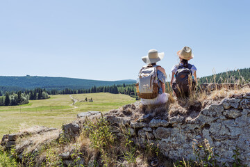 Mother and daughter hikers sitting on rocky viewpoint with backpacks, looking over sunny mountain meadow and forest, family travel rest and bonding in nature with panoramic landscape and copy space