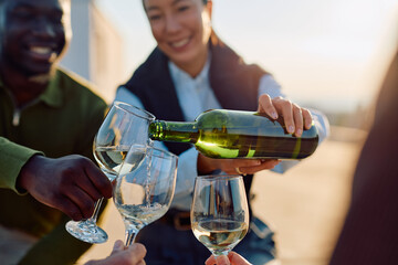 Friends on a rooftop at golden hour raising and pouring glasses of wine, smiling and celebrating together at sunset
