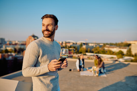 Smiling man enjoying a rooftop party with wine and phone, friends nearby. Urban lifestyle and relaxation concept