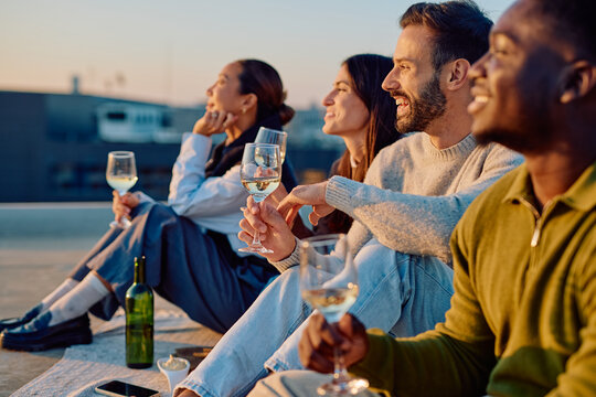 Diverse friends enjoying drinks and conversation during a rooftop party at sunset, embracing companionship and happiness - Powered by Adobe