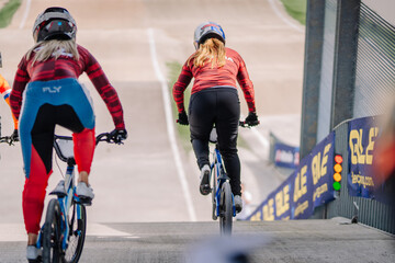 Fototapeta premium Valmiera, Latvia - July 12, 2025: Female BMX riders in colorful jerseys preparing to race on a professional track with vibrant surroundings