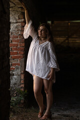 Naklejka premium Young woman with long hair wearing a white oversized shirt poses in an abandoned building, showcasing a blend of fashion and urban decay in a captivating atmosphere
