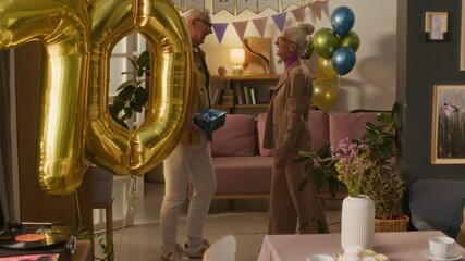 Excited senior woman warmly greeting friends showing up in her living room with birthday presents and inviting them to table