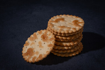 Stack of golden scalloped savoury crackers biscuits with one leaning, on dark surface.