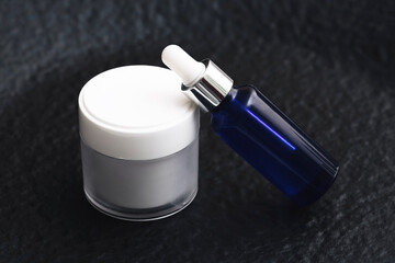 Cosmetic cream jar with blue glass serum bottle on dark background. Skincare and beauty product presentation. Minimal aesthetic for natural face treatment.