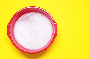 Red plastic basin with water and foam on yellow background, top view. Space for text