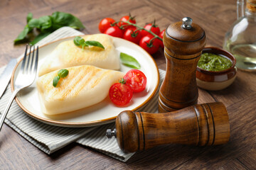 Salt, pepper mills, pieces of grilled squid, tomatoes, basil and sauce on wooden table, closeup