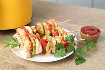 Tasty skewers with chicken, vegetables, parsley, sauce and air fryer on wooden table indoors, closeup