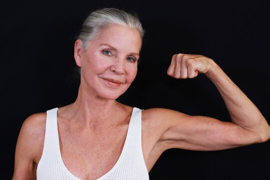 Fit strong attractive older woman flexing biceps with one arm, smiling looking at camera gray hair pulled back 