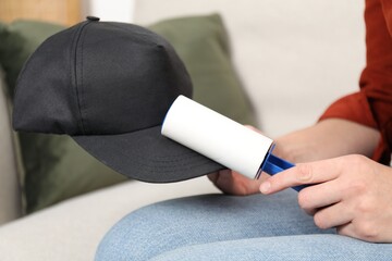 Woman cleaning black baseball hat with lint roller at home, closeup