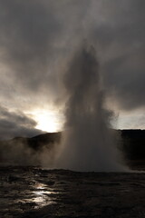 A powerful geyser erupts with a towering jet of water and steam, set against a dramatic sky with dark clouds and a glowing sun partially obscured by mist.