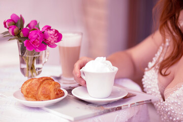 Close-up of a woman’s hand holding a cup of cappuccino with whipped cream at an outdoor cafe table with croissant and pink peonies in vase