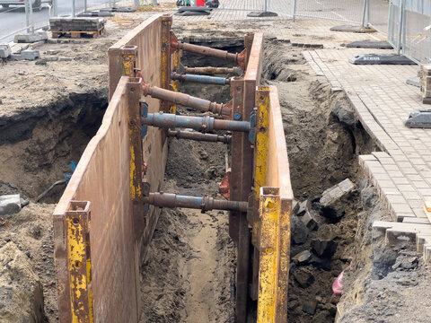 Busy urban construction site with a deep excavated trench, securely supported by a heavy-duty metal shoring system, for essential underground utility line installation