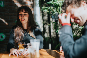 A woman smiles, sitting at a cafe table with a drink, as a man laughs energetically under the warm sunlight, creating a pleasant and relaxed atmosphere of connection.