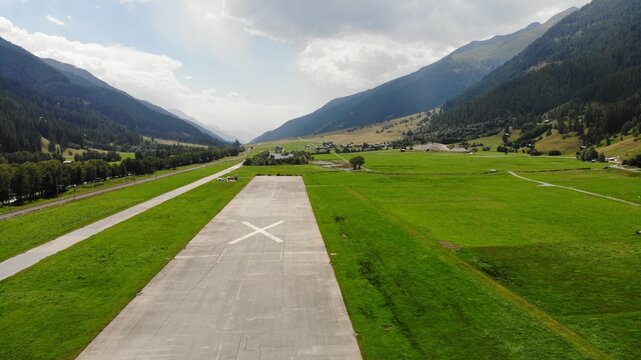 A small airstrip marked with a prominent white "X" indicating closure, nestled in a lush green valley surrounded by mountainous terrain