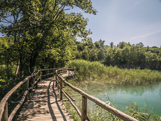 Winding wooden boardwalk follows a turquoise lake bordered by reeds and trees under a bright blue sky in Plitvice Lakes National Park.