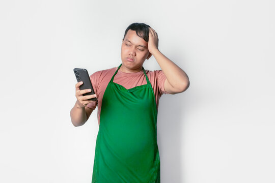 Asian man wearing green apron holding his head with confused expression and looking at his cell phone