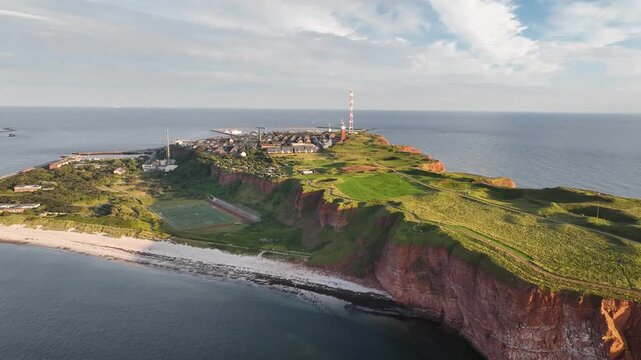 Aerial footage of Helgoland, Germany with red sandstone cliffs, green plateau, sandy beach, island&rsquo;s sports field and island village with harbor facilities and lighthouse. 
