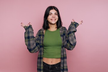 Young woman smiling with open arms against a pink background, wearing a green top and plaid jacket