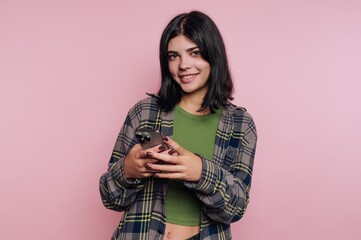 Young woman smiling while holding smartphone in front of pink background during casual indoor...